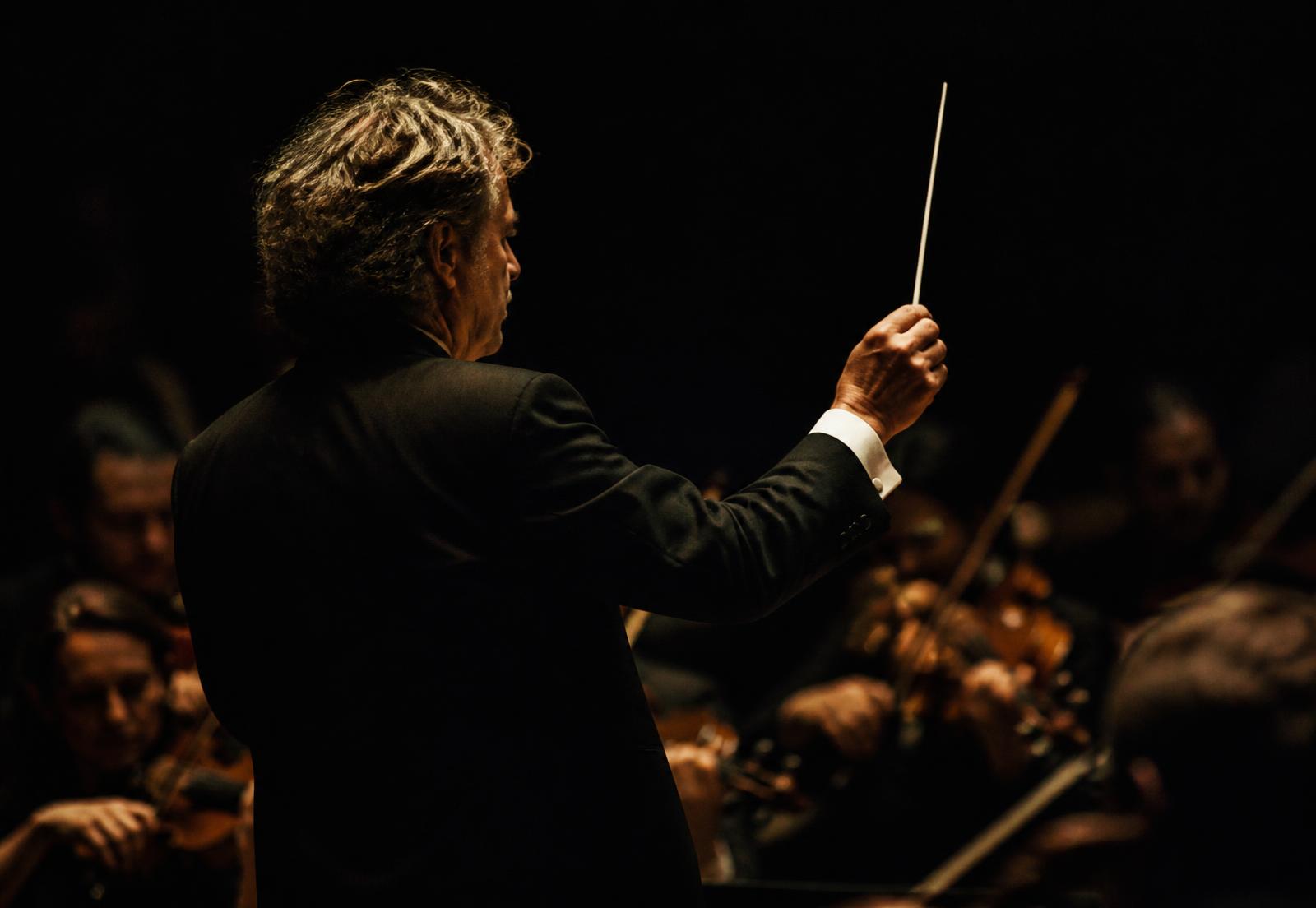 A conductor raising the baton before a shadowed orchestra